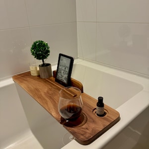 May include: A wooden bathtub tray with a wine glass, a small bottle, and a digital reader. The tray also holds a small potted plant and a candle. The digital reader displays the word "Wedding". The tray is placed across a white bathtub.
