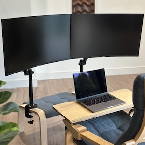 May include: A modern home office setup featuring two large black computer monitors mounted on adjustable stands. A laptop sits on a wooden desk attached to a gray upholstered chair. A matching footrest is also visible.