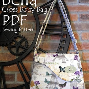 May include: A white and beige cross body bag with a floral and Eiffel Tower print. The bag has a silver clasp and a long strap. The bag is hanging on a black clock with gears. The text "Bella Cross Body Bag PDF Sewing Pattern" is visible in the image.