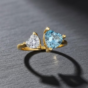 Może przedstawiać: Złoty pierścionek z dwoma kamieniami szlachetnymi w kształcie serca. Jeden jest przezroczysty, drugi jasnoniebieski. Pierścionek ma konstrukcję z rozciętym trzonkiem, z każdym kamieniem osadzonym po przeciwnych stronach obrączki. Pierścionek jest prezentowany na ciemnoszarej powierzchni.