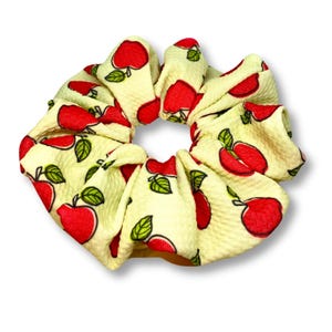 May include: A yellow scrunchie with a red apple pattern. The scrunchie is made of a soft, textured fabric.