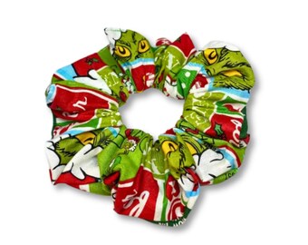 Puede incluir: Una goma para el pelo de tela con un diseño festivo en verde, rojo y blanco. El patrón incluye personajes de dibujos animados y texto, creando un accesorio temático de vacaciones. La goma para el pelo está diseñada para sujetar el cabello.