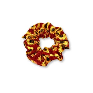May include: A scrunchie with a sunflower pattern. The fabric is a deep red with yellow sunflowers and black accents. The scrunchie is circular and has a gathered appearance.