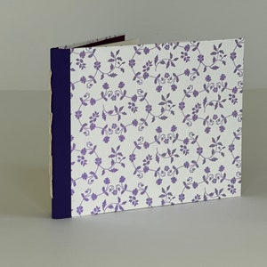 May include: A small, white notebook with a purple floral pattern on the cover. The notebook has a dark blue spine and is bound with a simple stitch.