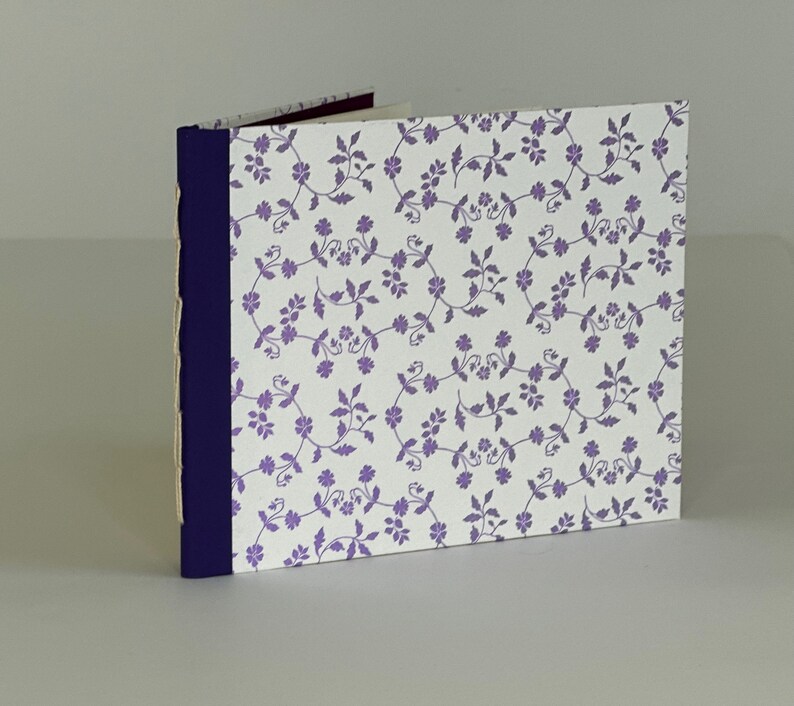 May include: A small, white notebook with a purple floral pattern on the cover. The notebook has a dark blue spine and is bound with a simple stitch.