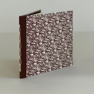 May include: A brown fabric-covered journal with a white swirling pattern. The journal has a white cord sewn along the spine.