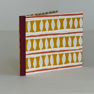 May include: A small, burgundy-colored book with a white and yellow patterned cover. The pattern features repeating white ovals on a yellow background with thin red lines.