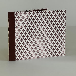 May include: A small, brown, handmade book with a white cover featuring a repeating pattern of dark brown fleur-de-lis.