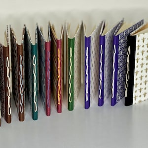 May include: A collection of 15 small, handmade books with various patterned covers and colorful stitching along the spine. The books are arranged in a row, with the spines facing the viewer.