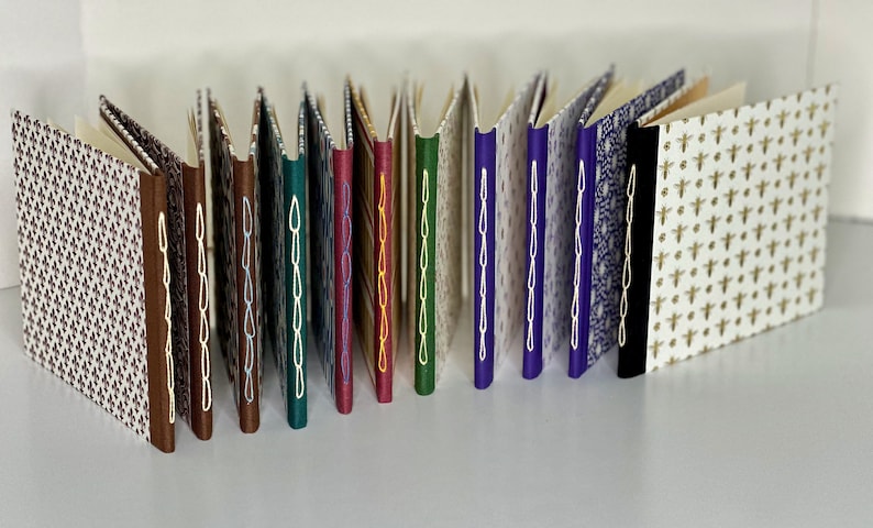 May include: A collection of 15 small, handmade books with various patterned covers and colorful stitching along the spine. The books are arranged in a row, with the spines facing the viewer.