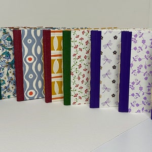 May include: A row of 16 small, handmade journals with various patterned covers. The covers feature different colors and designs, including floral, geometric, and abstract patterns. The journals are bound with colorful ribbon spines.