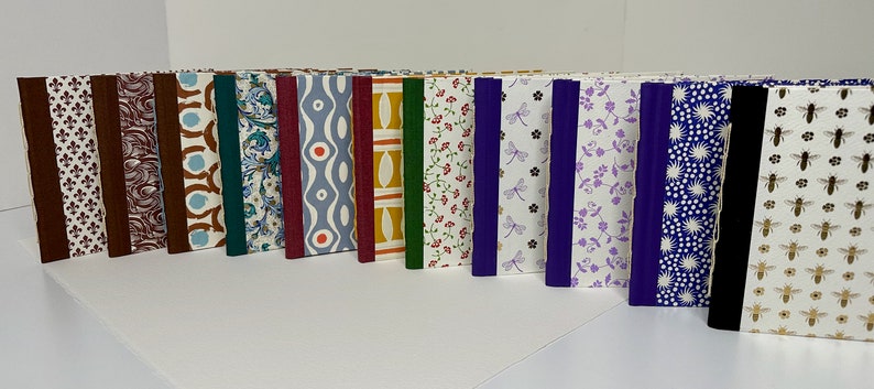 May include: A row of 16 small, handmade journals with various patterned covers. The covers feature different colors and designs, including floral, geometric, and abstract patterns. The journals are bound with colorful ribbon spines.