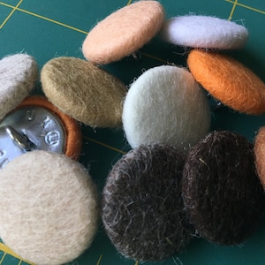 May include: A selection of felt buttons in assorted colours and shapes. The buttons are in shades of beige, white, orange, brown, and tan. Each button has a metal shank on the back for sewing onto fabric.