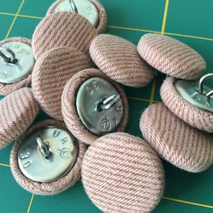 May include: A close-up of a pile of pink fabric covered buttons with metal shanks and engraved numbers. The buttons are arranged in a random pattern.