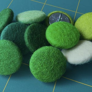 May include: A collection of green and white felt buttons, some with a textured surface. The buttons are arranged on a blue and yellow grid.