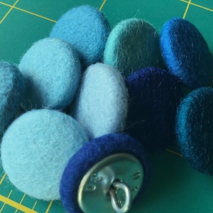 Könnte beinhalten: Eine Sammlung von blauen und türkisfarbenen Filzknöpfen in verschiedenen Schattierungen. Die Knöpfe sind rund und haben eine flauschige Textur.