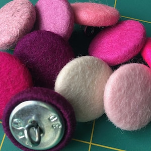 Pode incluir: Uma coleção de botões redondos de feltro em vários tons de rosa, roxo e branco. Os botões estão dispostos em um grupo sobre uma superfície verde.
