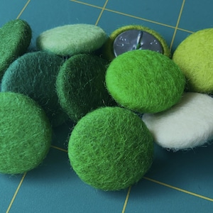 Può includere: Una collezione di bottoni in feltro verde e bianco, alcuni con una superficie testurizzata. I bottoni sono disposti su una griglia blu e gialla.