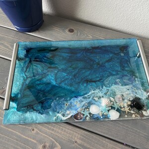 May include: Rectangular resin tray with a seascape design. The tray features shades of blue and white, with embedded seashells and a wave-like pattern. It has silver handles on each end.