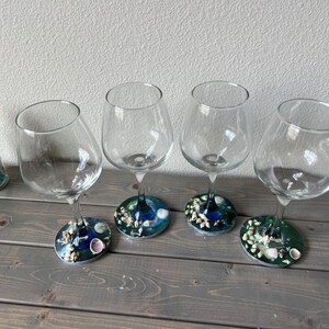 May include: Four clear wine glasses with decorative bases featuring a blue and green design with seashells. A small glass jar with a cork stopper and sea-themed contents sits to the left. The glasses are on a wooden surface.