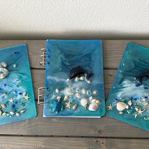 May include: Three ocean-themed notebooks with blue resin covers. Each notebook features a different design: a dolphin, a turtle, and the letter "S". Embellishments include seashells, starfish, and blue glass beads. The notebooks are on a wooden surface.