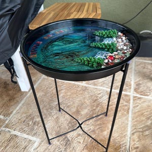 May include: A black metal side table with a round tray top. The tray features a miniature landscape with a blue and green water effect, miniature trees, and small rocks. The table has a black, angled leg design.