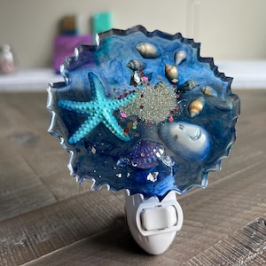 May include: A decorative night light with a blue resin design, a turquoise starfish, seashells, and glitter. The night light has a white base with an on/off switch. Ideal for a coastal or beach-themed room, it measures approximately 10 cm in diameter.