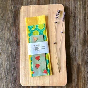 May include: A set of three reusable food wraps with a watermelon and lime print. The wraps are made of beeswax, jojoba oil, and tree resin. The wraps are on a wooden cutting board with a sprig of lavender.