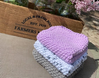May include: A wooden crate with the text "LOCAL & FRESH FOOD" holds three glass jars filled with lilac flowers. Stacked in front are three knitted washcloths in grey, light blue, and lavender. The scene is set outdoors on a sunny day.
