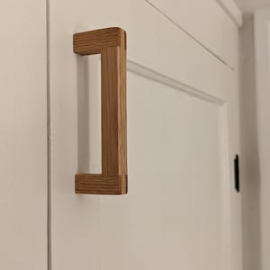 May include: A light wood cabinet door handle with a rectangular shape. The handle is made of natural wood and has a simple, minimalist design.