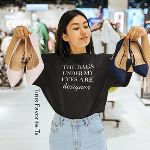 May include: A woman wearing a black t-shirt with the text "THE BAGS UNDER MY EYES ARE designer" in white lettering. She is holding two pairs of shoes, one pair is pink and the other is blue.