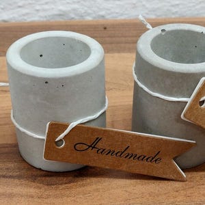 May include: Two small, gray concrete candle holders with a brown paper tag that says "Handmade".