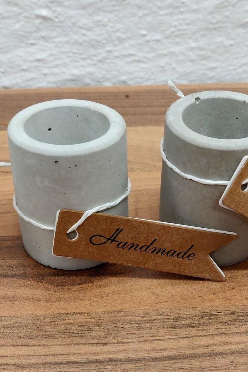 May include: Two small, gray concrete candle holders with a brown paper tag that says "Handmade".