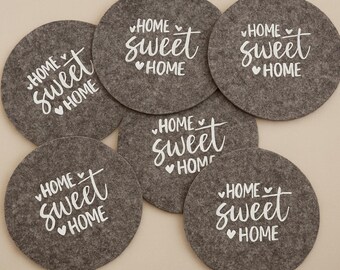 May include: A collection of gray felt coasters, each featuring the white text "HOME SWEET HOME" with a heart symbol. The coasters are round and arranged in a circular pattern, suitable for home decor.