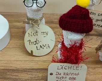 May include: Three white llama figurines with different accessories. The first llama is wearing a green hat and glasses and has a sign that says "Not my Prob - LAMA". The second llama is wearing a red knitted hat and scarf and has a sign that says "LACHLE! Du kannst nicht alle anspucken.". The third llama is wearing a straw hat and glasses and has a sign that says "Ein LA für gute RMA".
