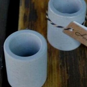 May include: Two small, light gray concrete cups with a rough texture. The cups are empty and appear to be handmade.
