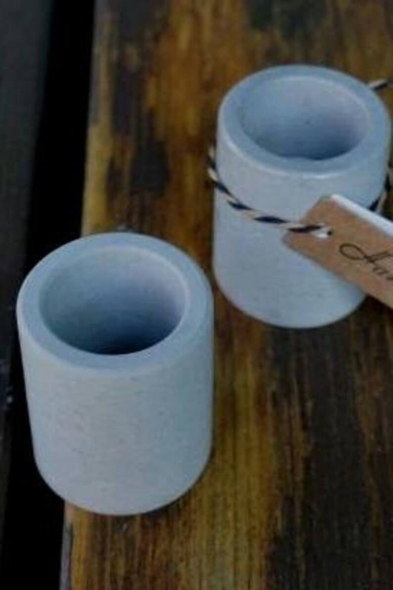 May include: Two small, light gray concrete cups with a rough texture. The cups are empty and appear to be handmade.