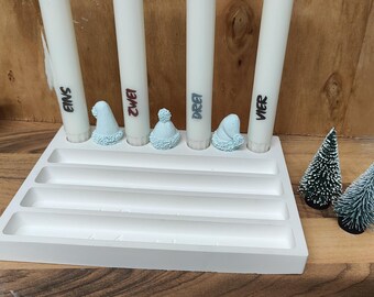 May include: A white Advent candle holder with four white candles labeled Eins, Zwei, Drei, and Vier. Each candle has a light blue hat. Two small green Christmas trees are on the right.