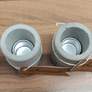 May include: Two gray concrete candle holders with a silver metal insert for a tea light. The holders are tied with twine and have a brown paper tag that says "Handmade".