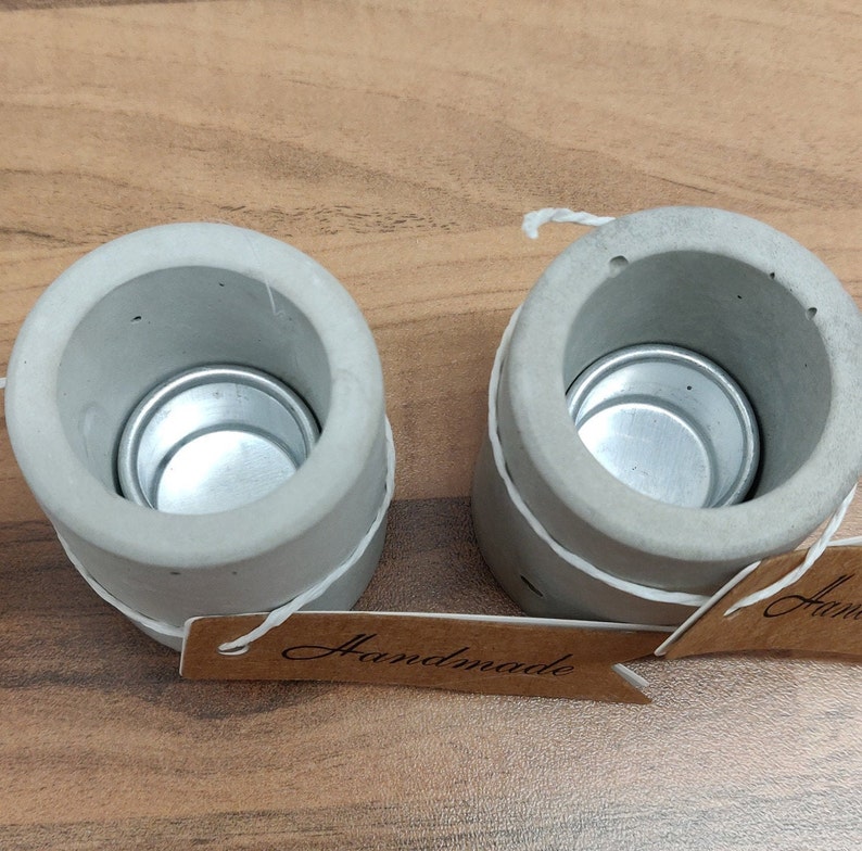 May include: Two gray concrete candle holders with a silver metal insert for a tea light. The holders are tied with twine and have a brown paper tag that says "Handmade".