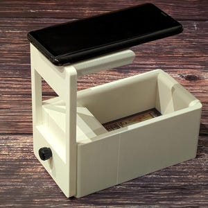 May include: White 3D printed phone stand with a slot for a phone and a drawer underneath. The phone is positioned above the drawer and is facing down.