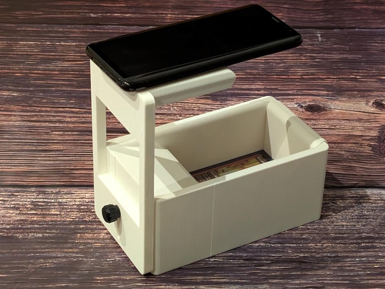 May include: White 3D printed phone stand with a slot for a phone and a drawer underneath. The phone is positioned above the drawer and is facing down.