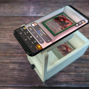 May include: A white phone stand with a black smartphone on top. The phone is displaying a mobile app with a card game interface. The app shows a card with a red and green design and the text "Forest  Lumberer".