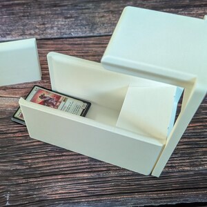 May include: White plastic card storage box with a sliding lid and a single card inside. The card is a Magic: The Gathering card with a picture of a creature on it.