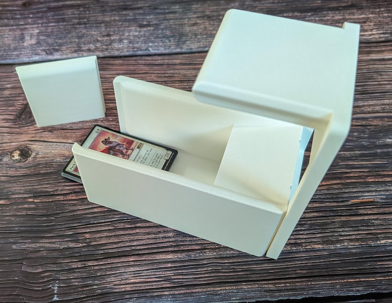 May include: White plastic card storage box with a sliding lid and a single card inside. The card is a Magic: The Gathering card with a picture of a creature on it.