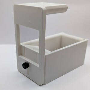 May include: White plastic 3D printed box with a small black knob on the side. The box has a removable top and a small opening on the front.