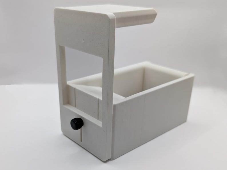 May include: White plastic 3D printed box with a small black knob on the side. The box has a removable top and a small opening on the front.