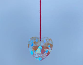 Puede incluir: Un colgante de corazón de cristal colgando de un hilo rojo. El corazón está facetado e iridiscente, reflejando colores arcoíris.