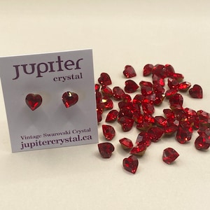 Könnte beinhalten: Ein Paar rote herzförmige Kristallohrringe mit goldenen Stiften. Die Ohrringe werden auf einer weißen Karte mit dem Text "jupiter crystal" und "Vintage Swarovski Crystal jupitercrystal.ca" gezeigt. Eine Streuung von roten herzförmigen Kristallen ist ebenfalls zu sehen.