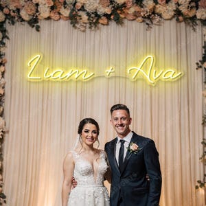 May include: A wedding photo featuring a couple posing in front of a floral backdrop and a neon sign that reads "Liam + Ava." The bride wears a white wedding dress, and the groom is in a black suit. The background includes a curtain and floral arrangements.